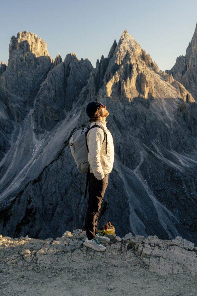 Fotografia przedstawia osobę stojącą na kamienistym wzniesieniu, na tle monumentalnych, ostrych górskich formacji. Ma na sobie jasną kurtkę, ciemne spodnie i plecak, a głowę unosi ku górze, jakby obserwowała szczyty. Światło padające z boku podkreśla fakturę skał i nadaje krajobrazowi surowy, monumentalny charakter.