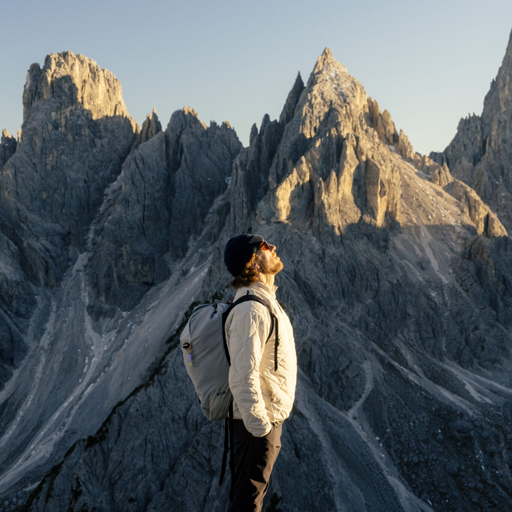 Osoba stojąca na górskim szlaku, z plecakiem, patrząca w górę na skaliste szczyty oświetlone niskim słońcem.